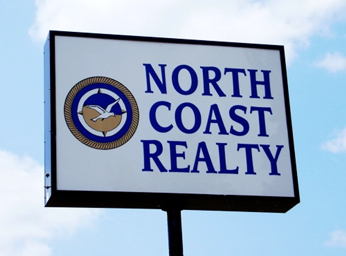 A double-sided white pole sign reading “North Coast Realty” against a blue sky